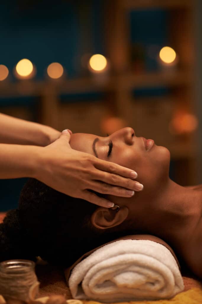 Massage relaxant visage et cuir chevelu : détente profonde Femme noire au visage détendu recevant un massage relaxant du cuir chevelu et du visage avec des mains expertes, lumière tamisée.